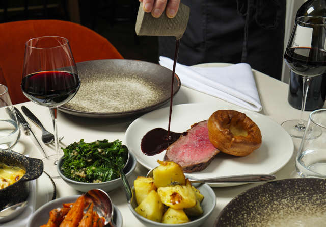 Elegant dinner at Virgin Hotels with roast beef, Yorkshire pudding, roasted vegetables, and red wine being poured on the plate.