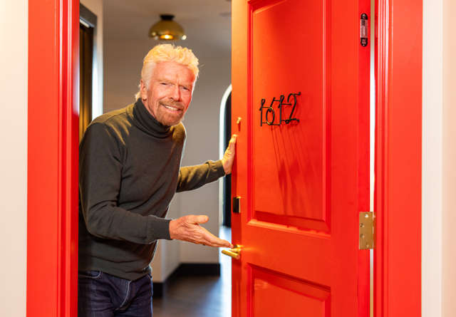 Richard Branson opening a bright red hotel room door, inviting guests into a warmly lit and stylish room at Virgin Hotels.