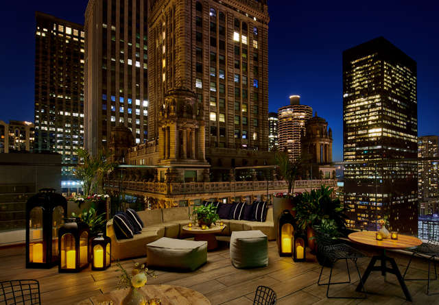 Nighttime view of the Cerise rooftop patio at Virgin Hotels, featuring cozy seating, ambient string lights, and a city skyline backdrop.